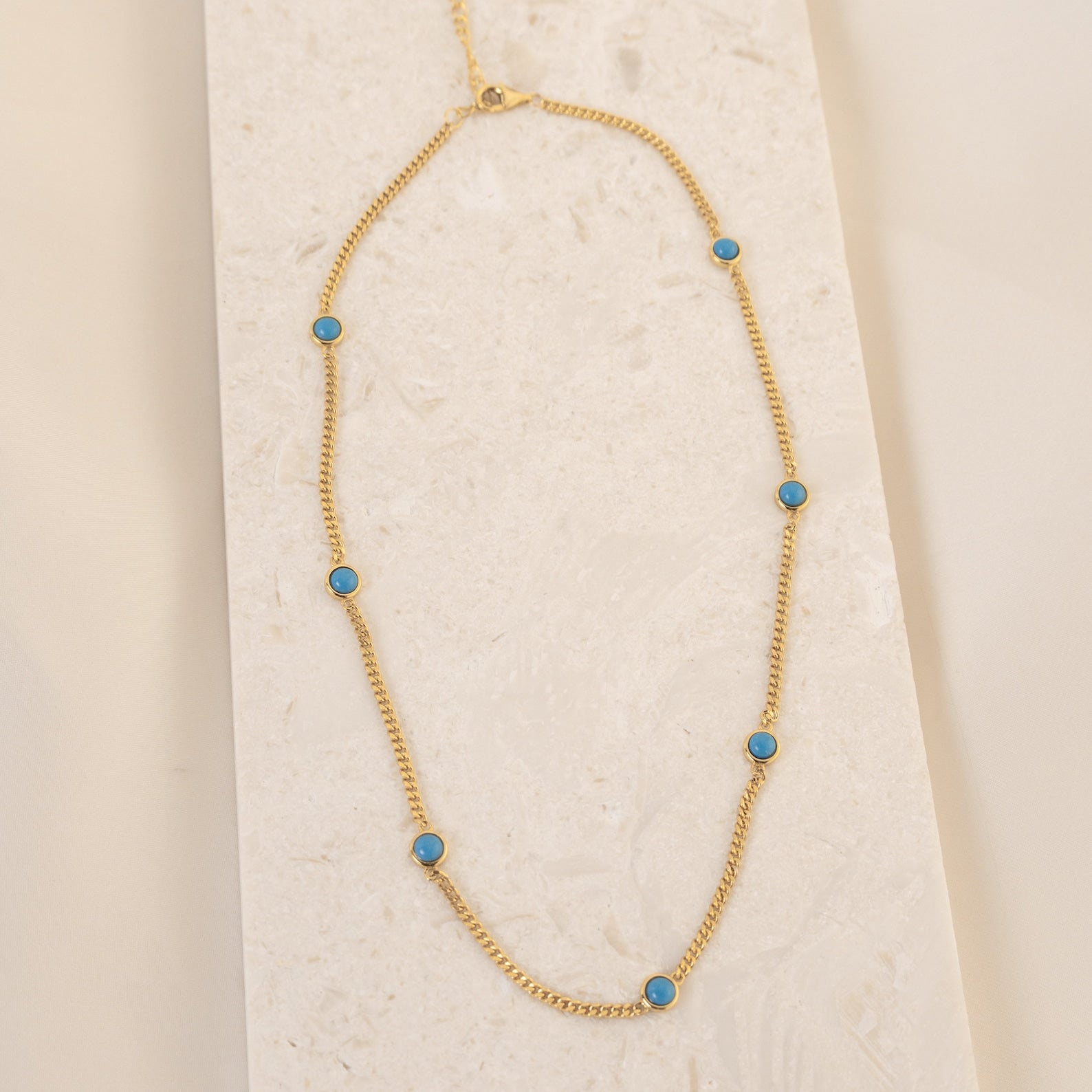 The Santorini Turquoise Choker features a gold chain with turquoise drops, beautifully displayed on a light textured surface.