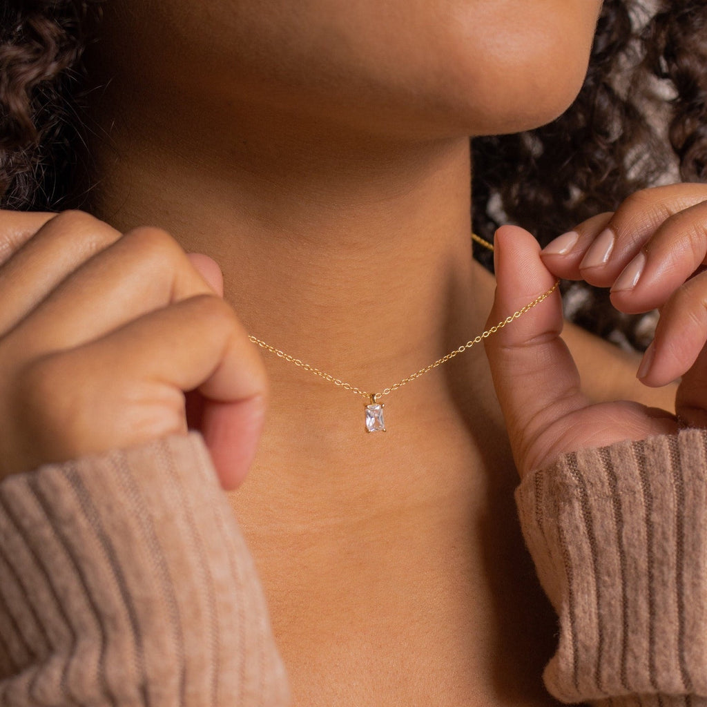 A person wearing the Sage Diamond Necklace with its small rectangular pendant, holding it near their neck and exuding timeless elegance.