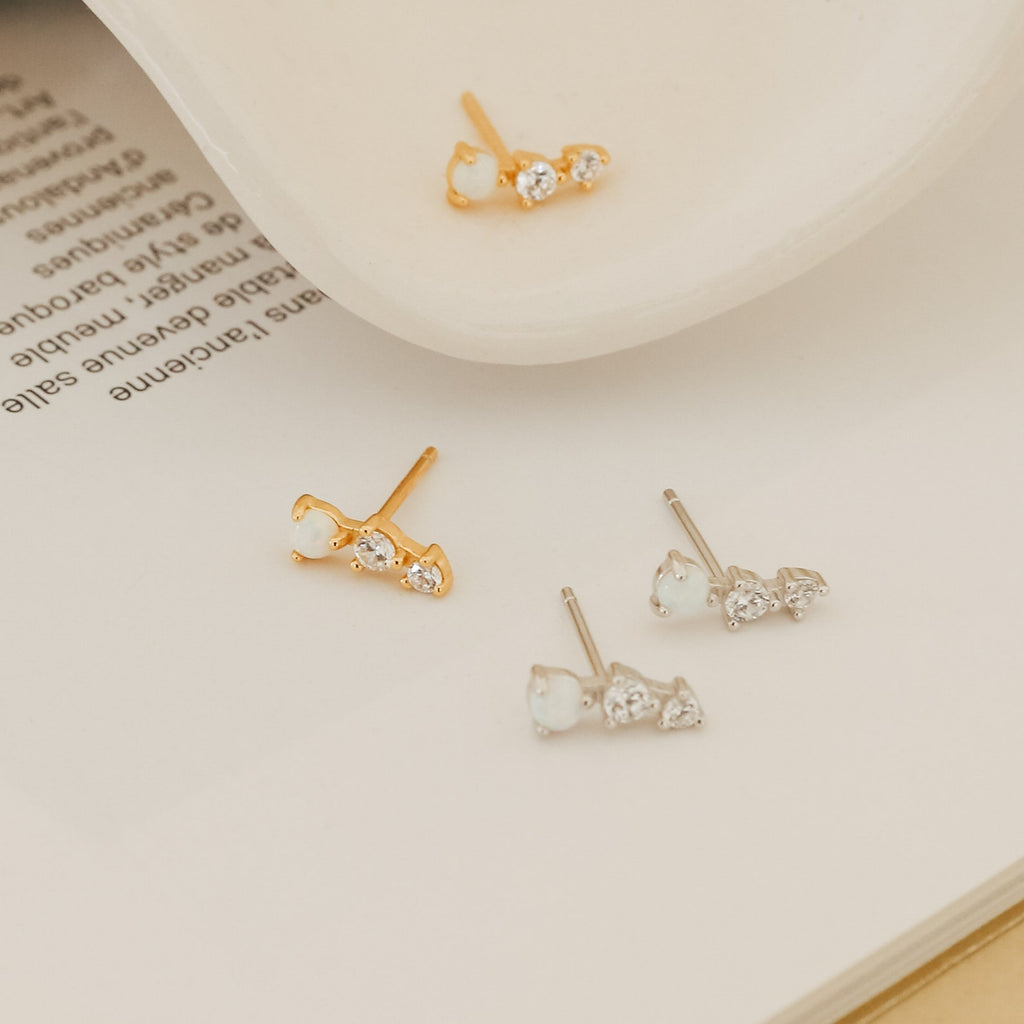 The Maeve Opal Studs, available in gold and silver with three opals per pair, are displayed on a book and a dish. These elegant earrings add a subtle statement to any look.