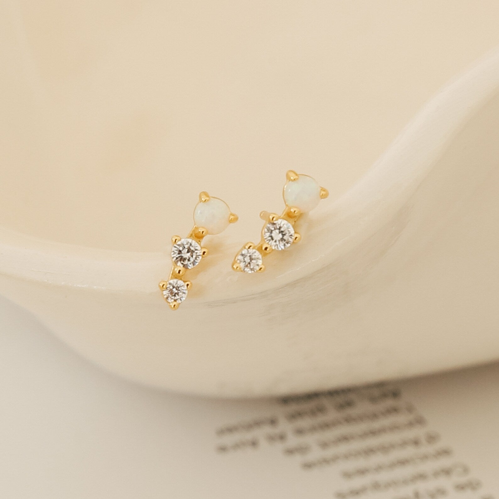Maeve Opal Studs: Gold statement earrings accented with two white stones and two clear crystals per earring, displayed on a cream ceramic dish.