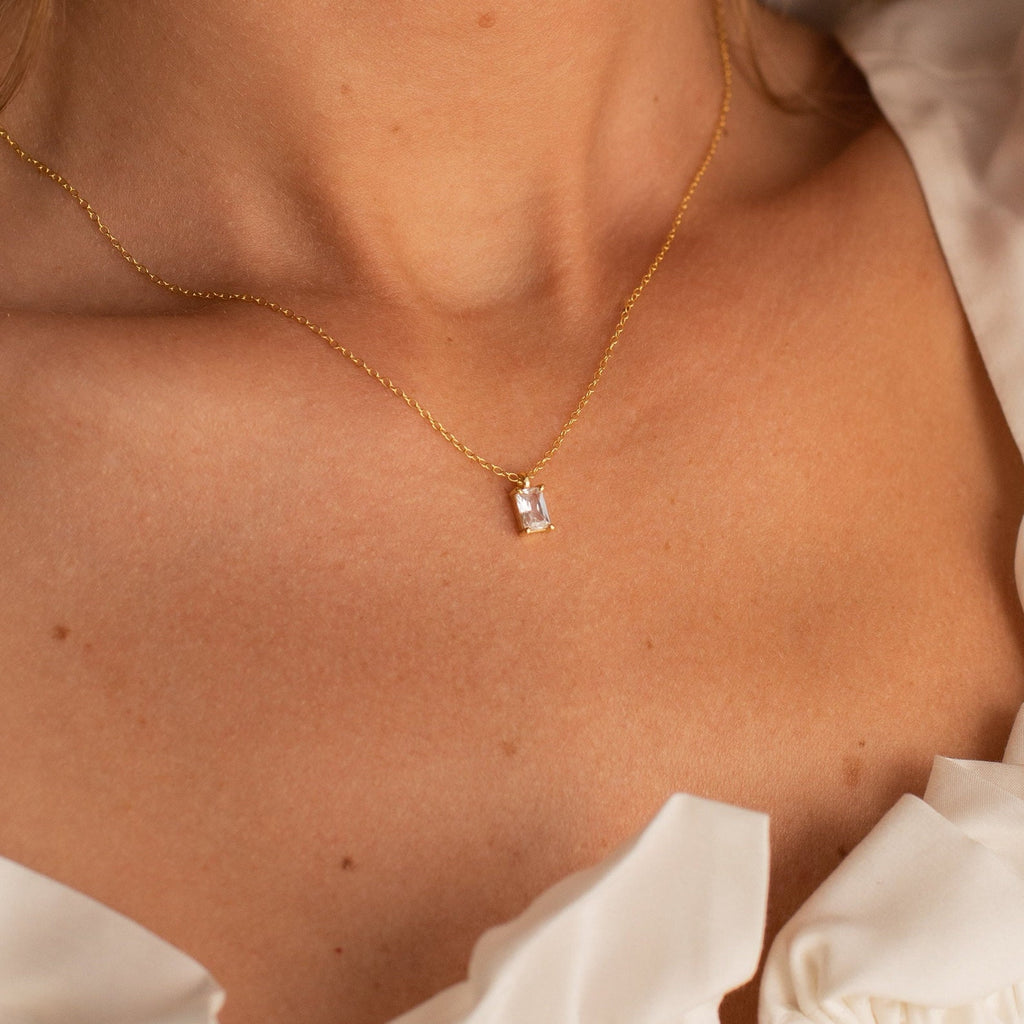Gold necklace featuring a rectangular emerald-cut crystal pendant, worn by a model in a white ruffled top, highlighting the clean, geometric lines and radiant clarity of the stone.