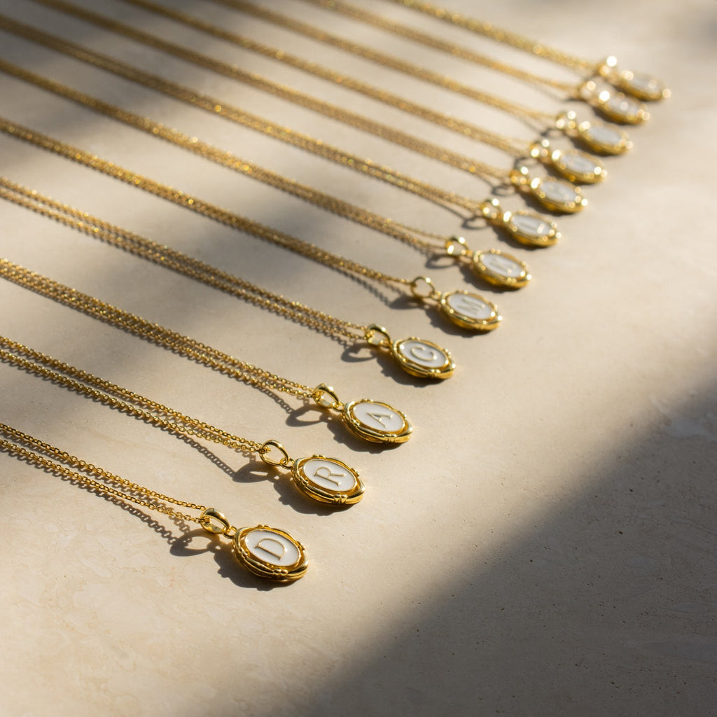 Initial Pendant Necklace, featuring a gold enamel finish and vintage-inspired design, is elegantly arranged in a row on a light surface and shimmers beautifully in sunlight.