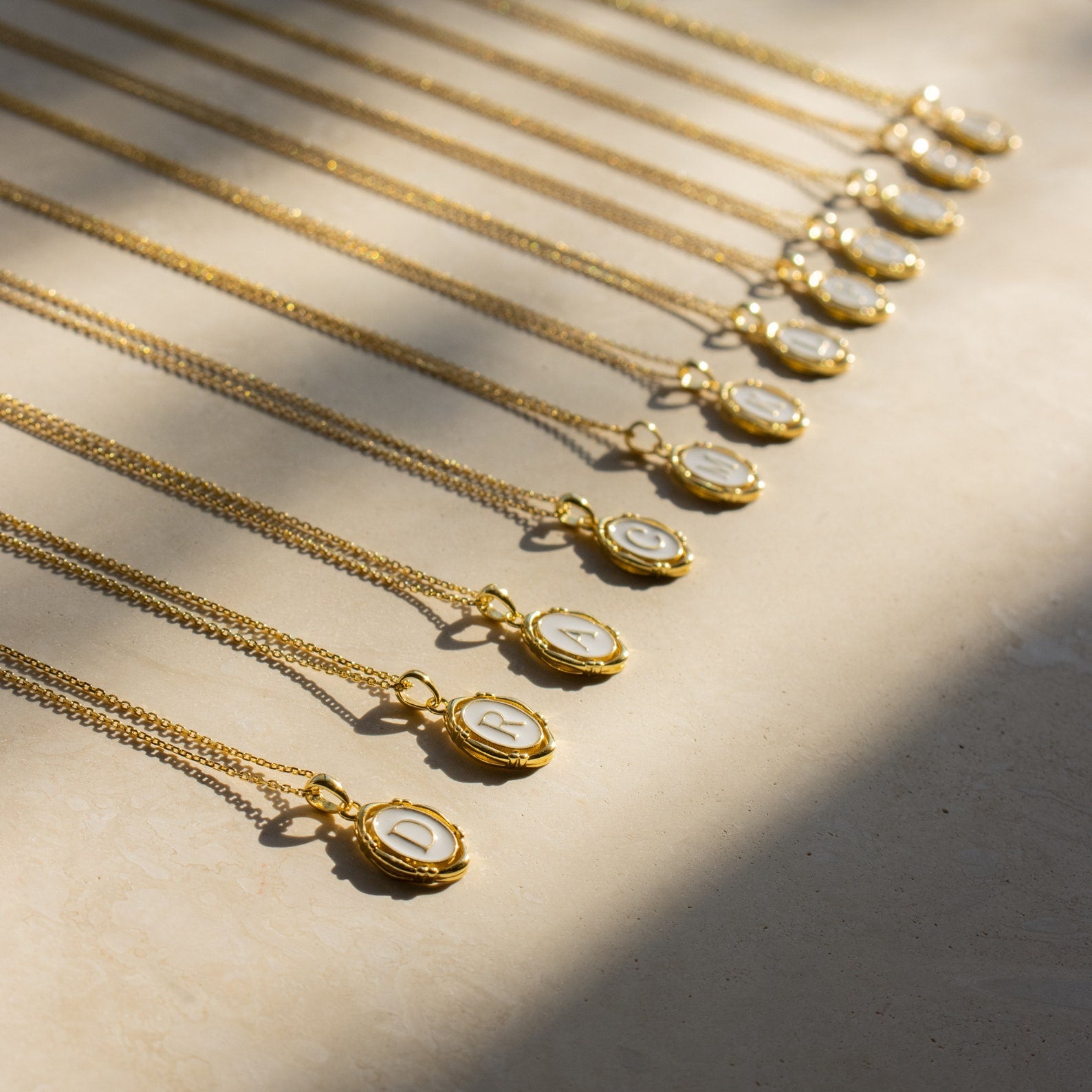 Initial Pendant Necklace, featuring a gold enamel finish and vintage-inspired design, is elegantly arranged in a row on a light surface and shimmers beautifully in sunlight.