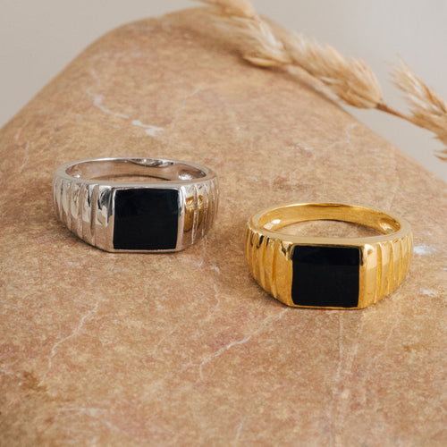 The Black Signet Ring features a bold rectangular black stone, available in both silver and gold finishes. Its unisex design makes it a striking accessory for any style, shown here on a textured stone surface.