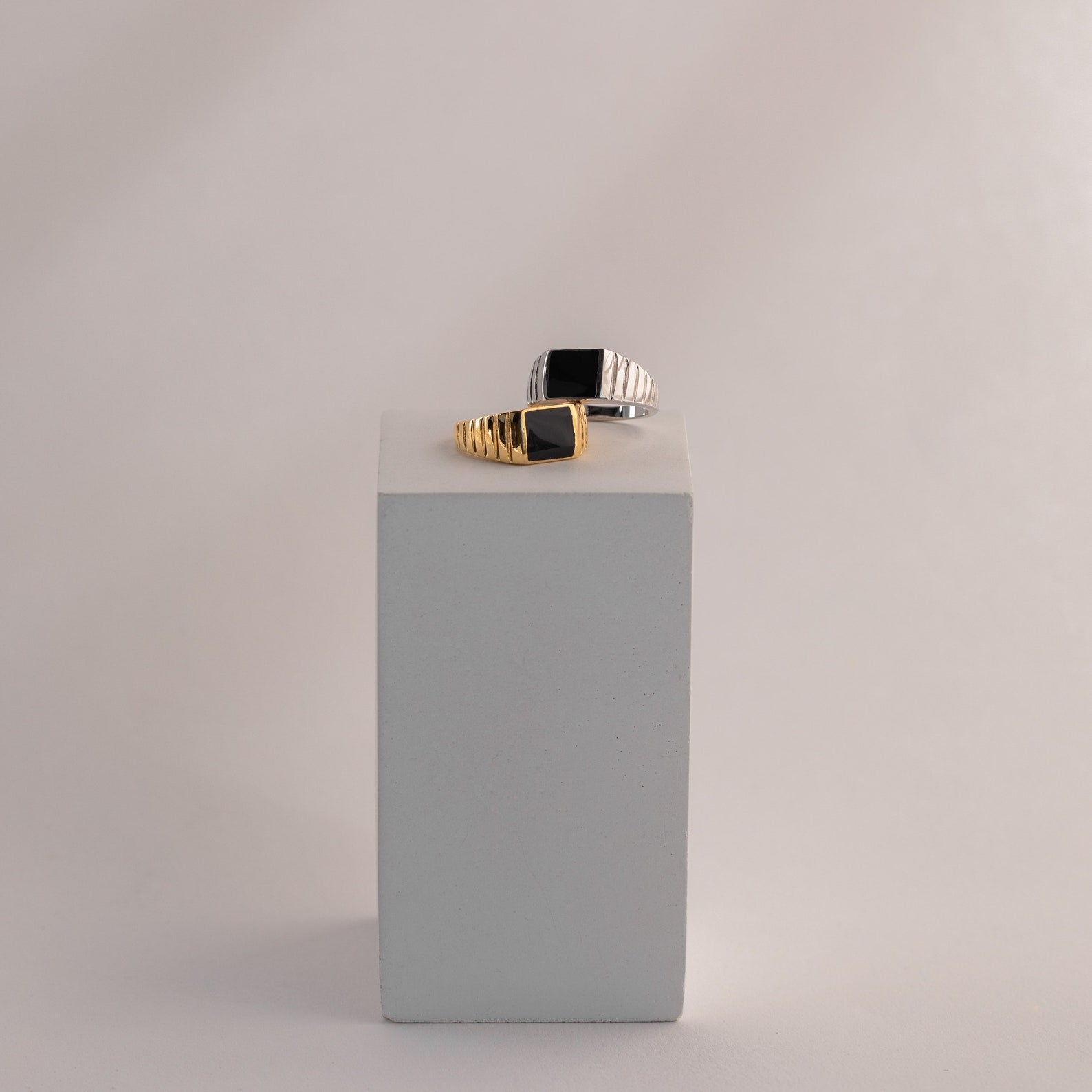 Two Black Signet Rings—square, unisex, one with gold and one with silver accents—are displayed on a minimalist gray block. These bold statement rings suit any style.
