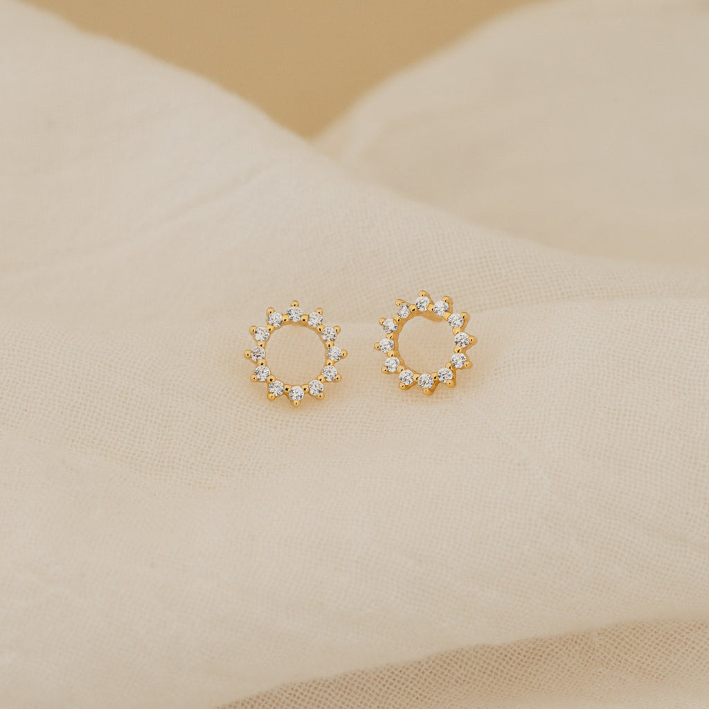 Helen Diamond Sun Studs—gold circular minimal earrings with small clear stones—are displayed on soft beige fabric. These dainty diamond studs add a subtle sparkle to any look.