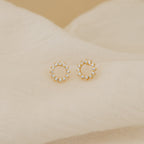 Helen Diamond Sun Studs—gold circular minimal earrings with small clear stones—are displayed on soft beige fabric. These dainty diamond studs add a subtle sparkle to any look.
