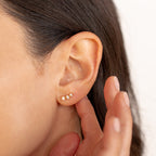 Close-up of a woman's ear with three small gold and pearl stud earrings, her hand touching her earlobe—perfect for pairing with statement earrings like the vibrant Maeve Opal Studs.