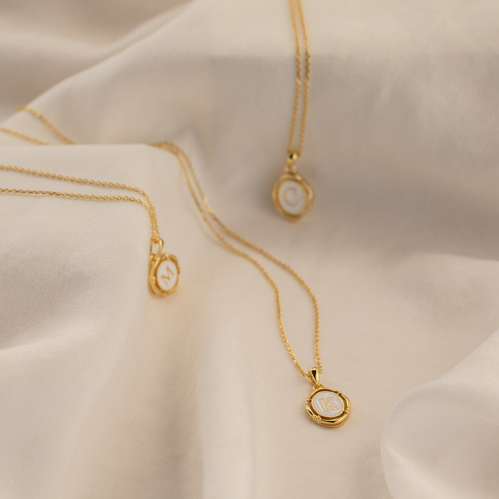 Three Initial Pendant Necklaces in 18K Gold, each featuring a round charm engraved with a C, M, or K, are displayed on soft fabric.
