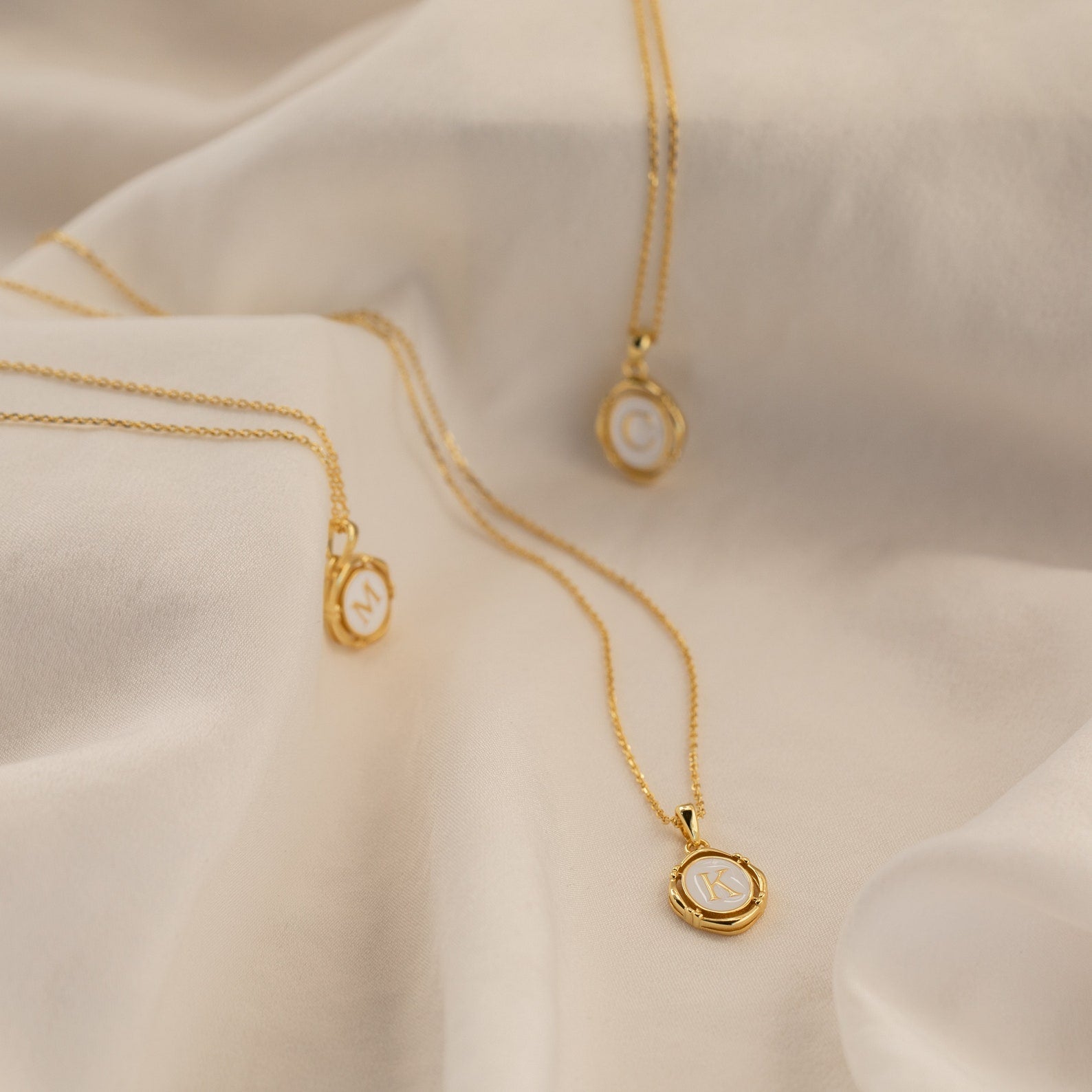Three Initial Pendant Necklaces in 18K Gold, each featuring a round charm engraved with a C, M, or K, are displayed on soft fabric.