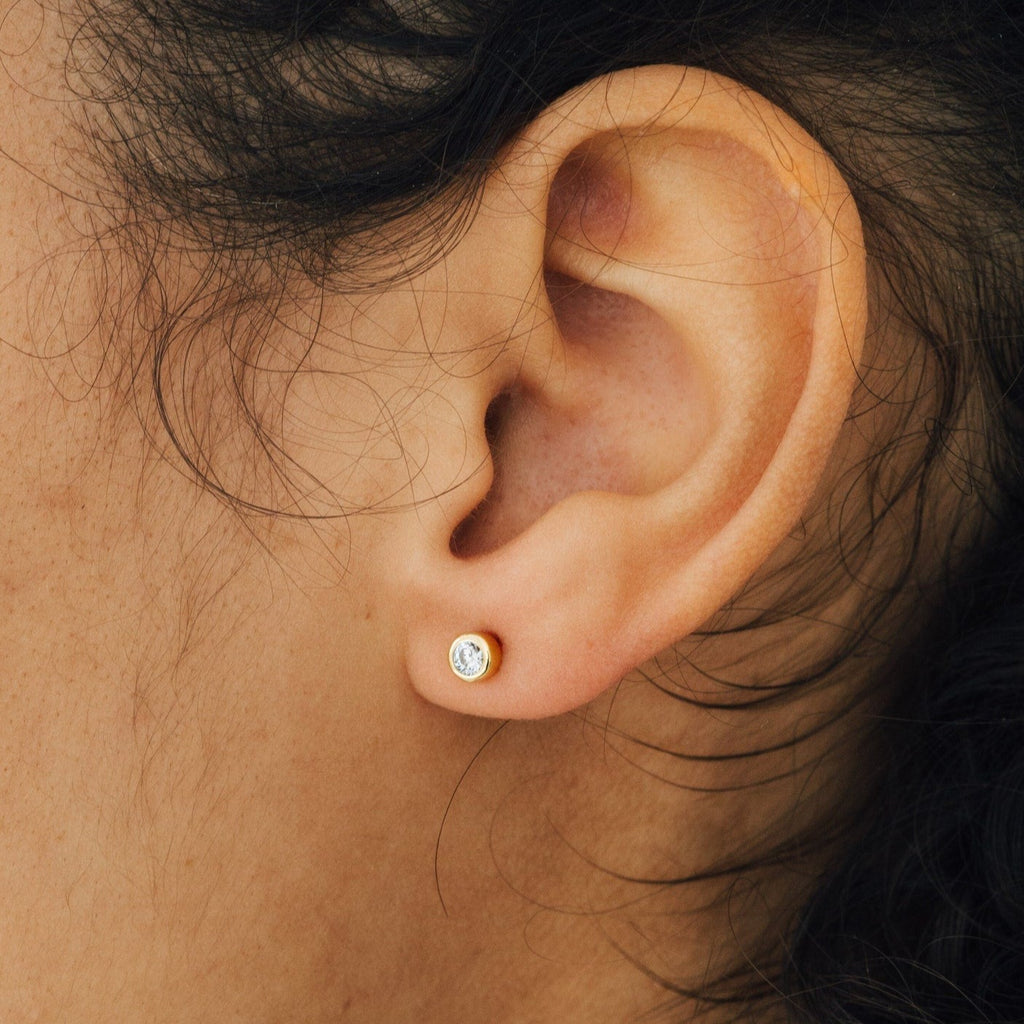 Close-up of a person’s ear wearing the Jane Diamond Studs—small gold earrings featuring a bezel set round brilliant diamond.