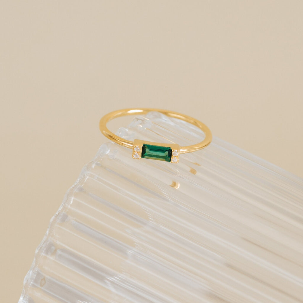 The Empire Emerald Ring features a rectangular green emerald accented with small diamonds, showcased elegantly on a clear ribbed surface.