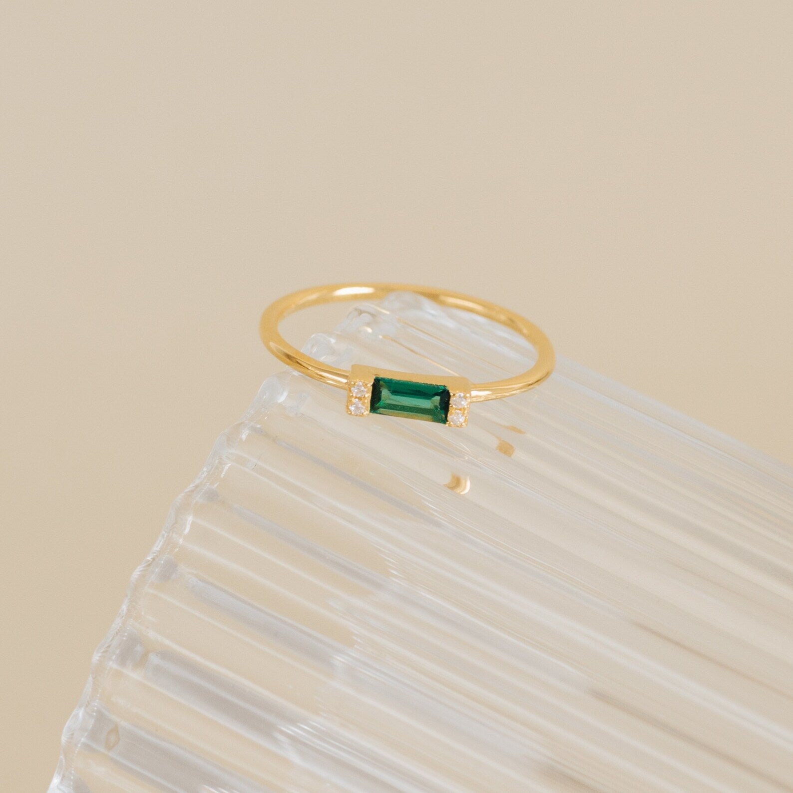 The Empire Emerald Ring features a rectangular green emerald accented with small diamonds, showcased elegantly on a clear ribbed surface.