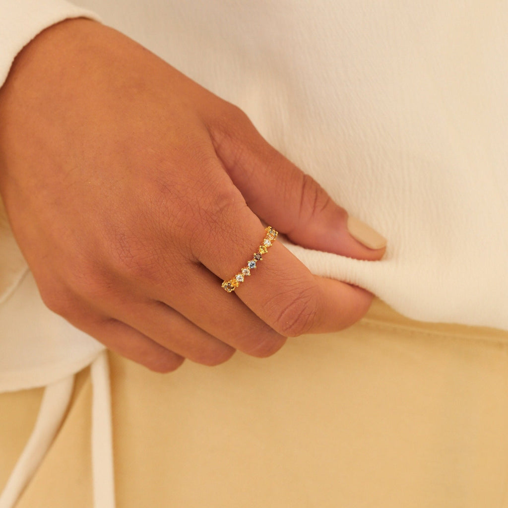 A manicured hand wearing the Rainbow Crystal Ring, adorned with pastel gemstones, rests on cream-colored fabric.