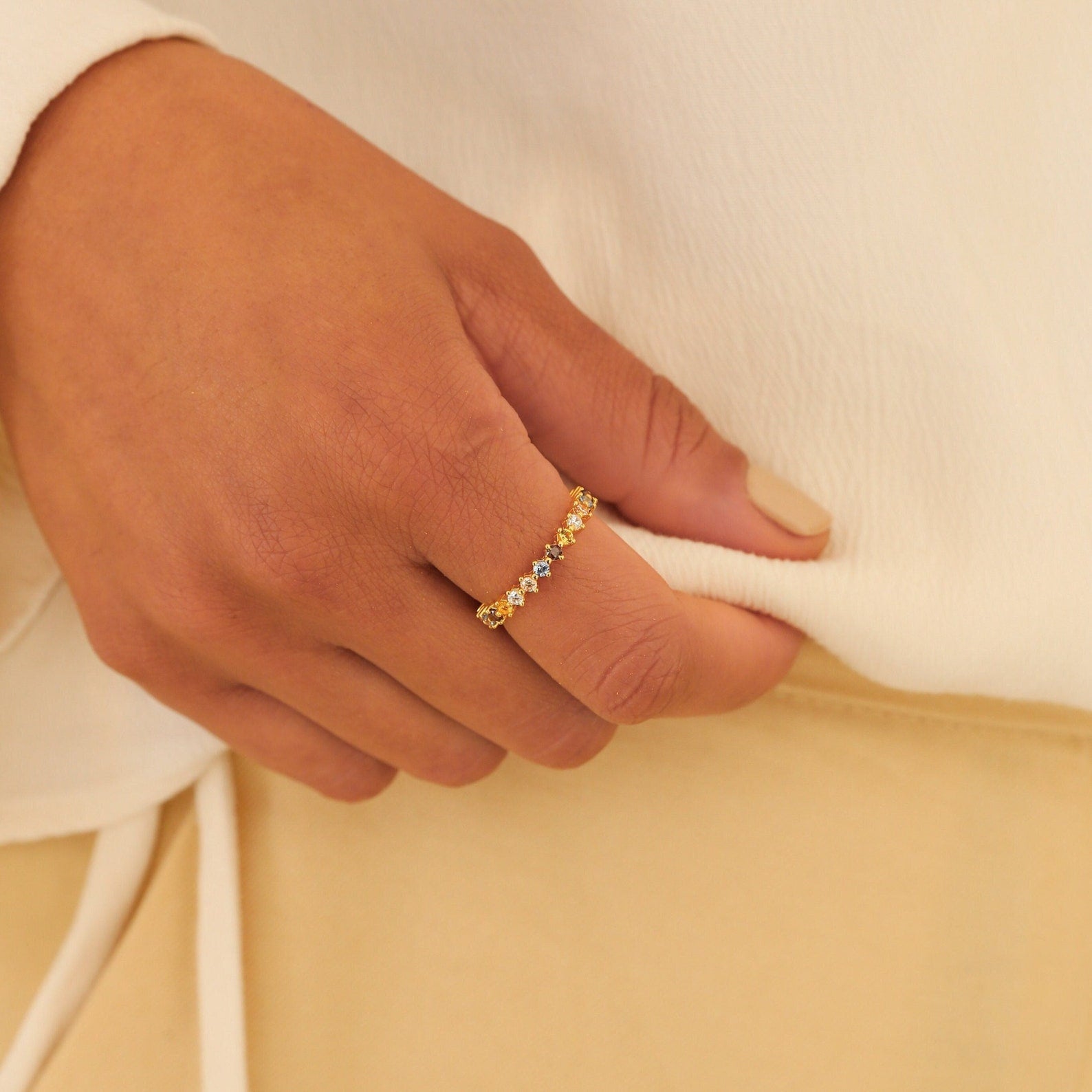 A manicured hand wearing the Rainbow Crystal Ring, adorned with pastel gemstones, rests on cream-colored fabric.