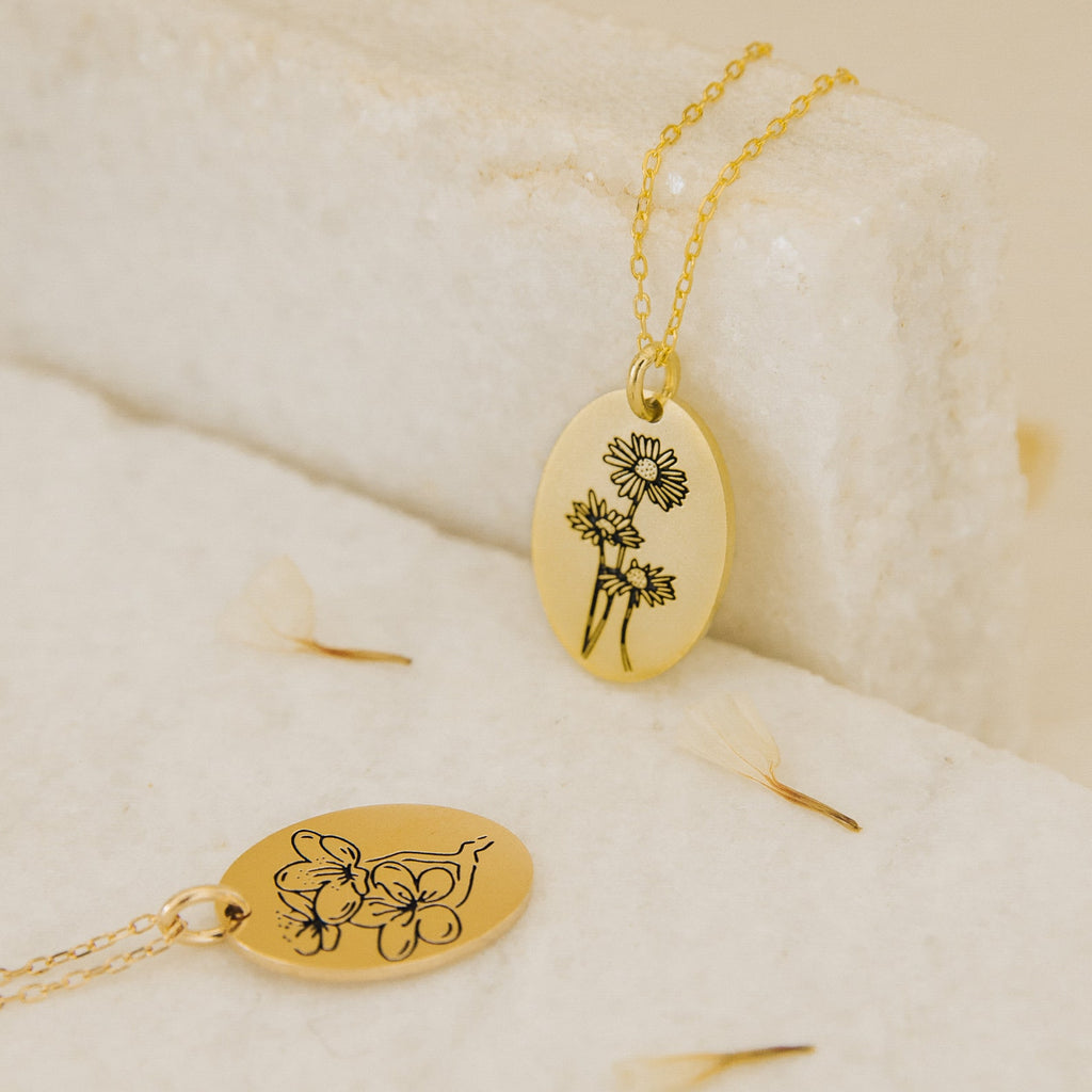 Two Oval Flower Necklaces with delicate floral engravings rest on a light, textured surface with scattered petals, showcasing unique flower jewelry.