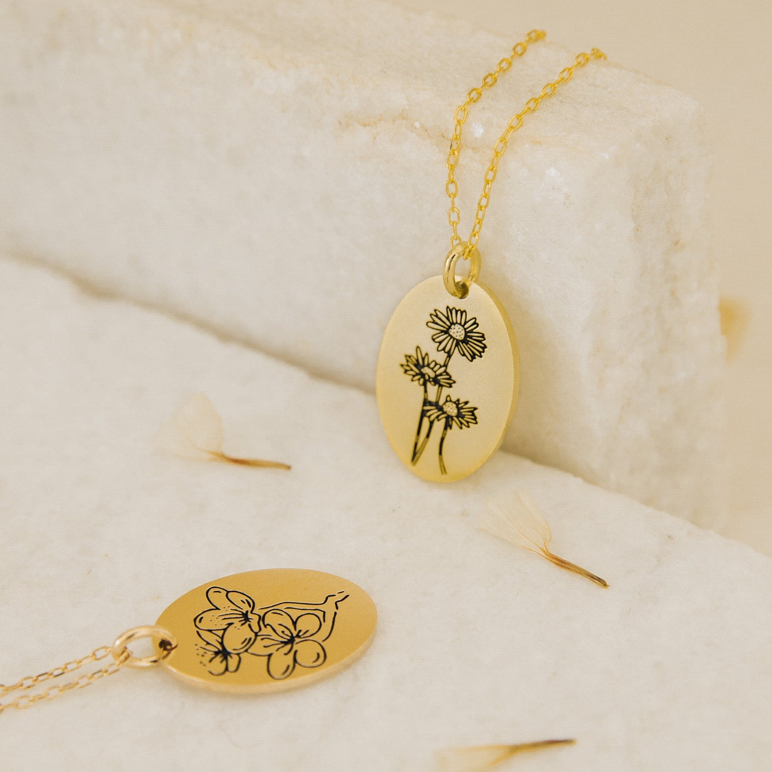 Two Oval Flower Necklaces with delicate floral engravings rest on a light, textured surface with scattered petals, showcasing unique flower jewelry.