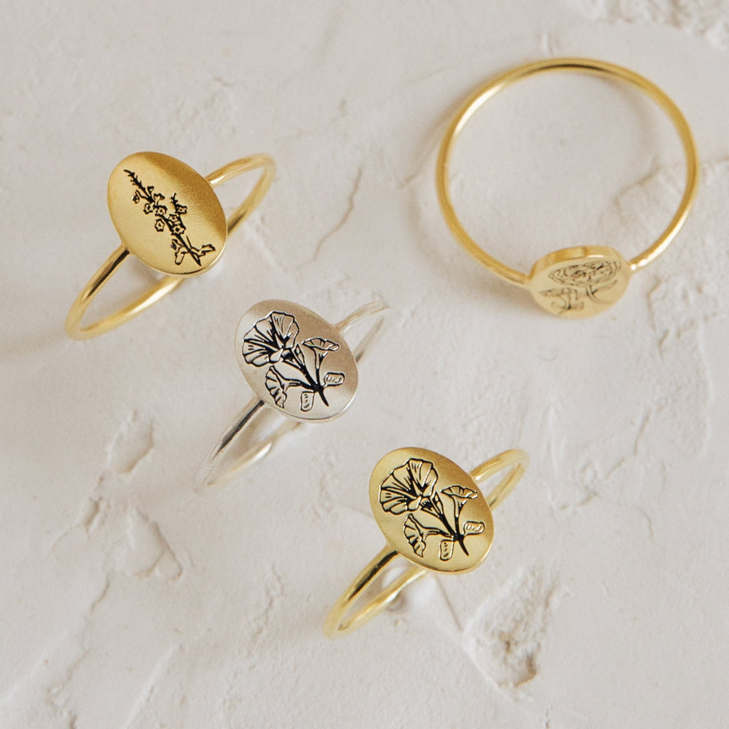 The Flower Oval Ring, available in gold and silver with engraved birth flowers, is displayed on a textured white surface—an ideal choice for personalized floral jewelry gifts.