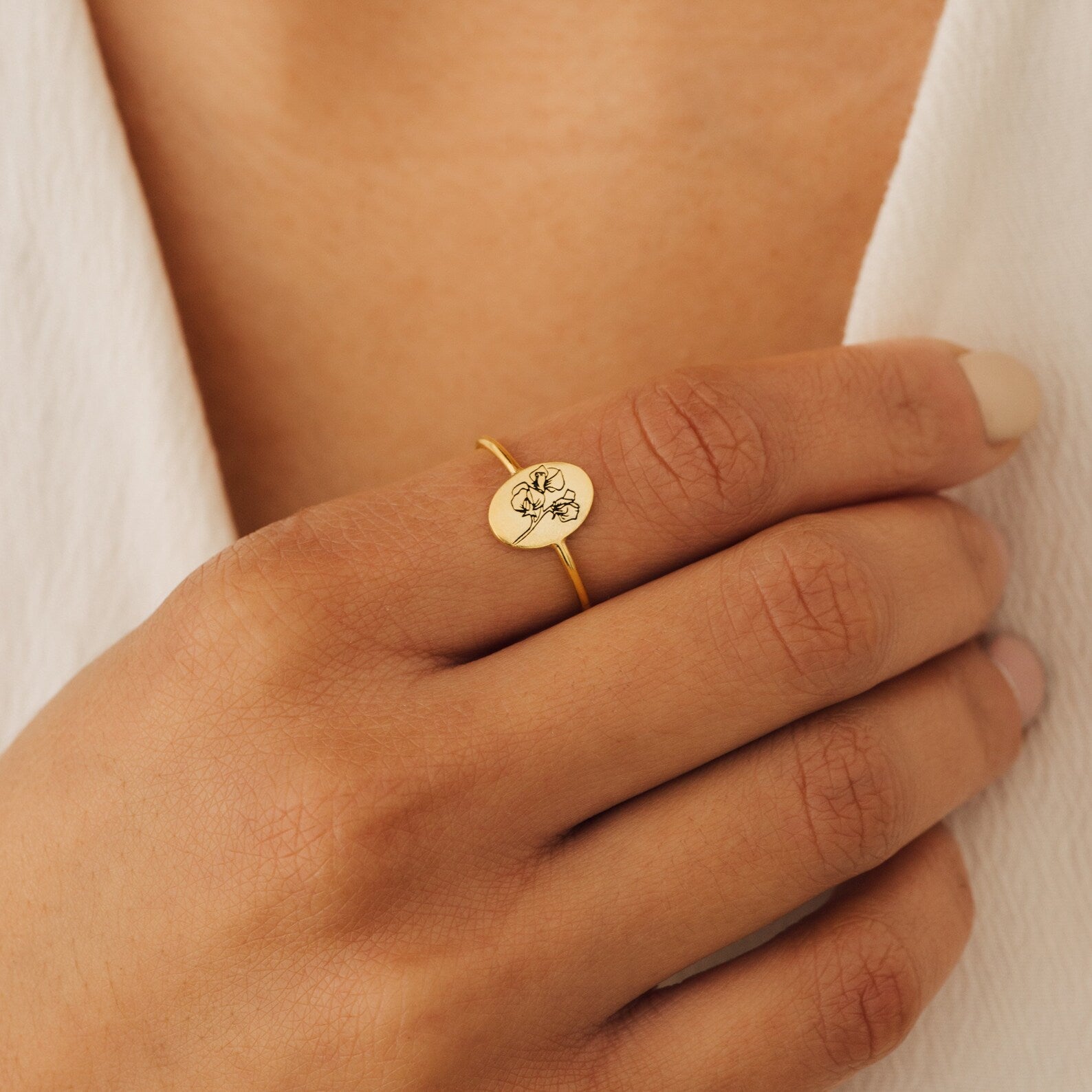 A hand wears the Flower Oval Ring, showcasing an engraved bloom design in gold, set against a white textured fabric background.