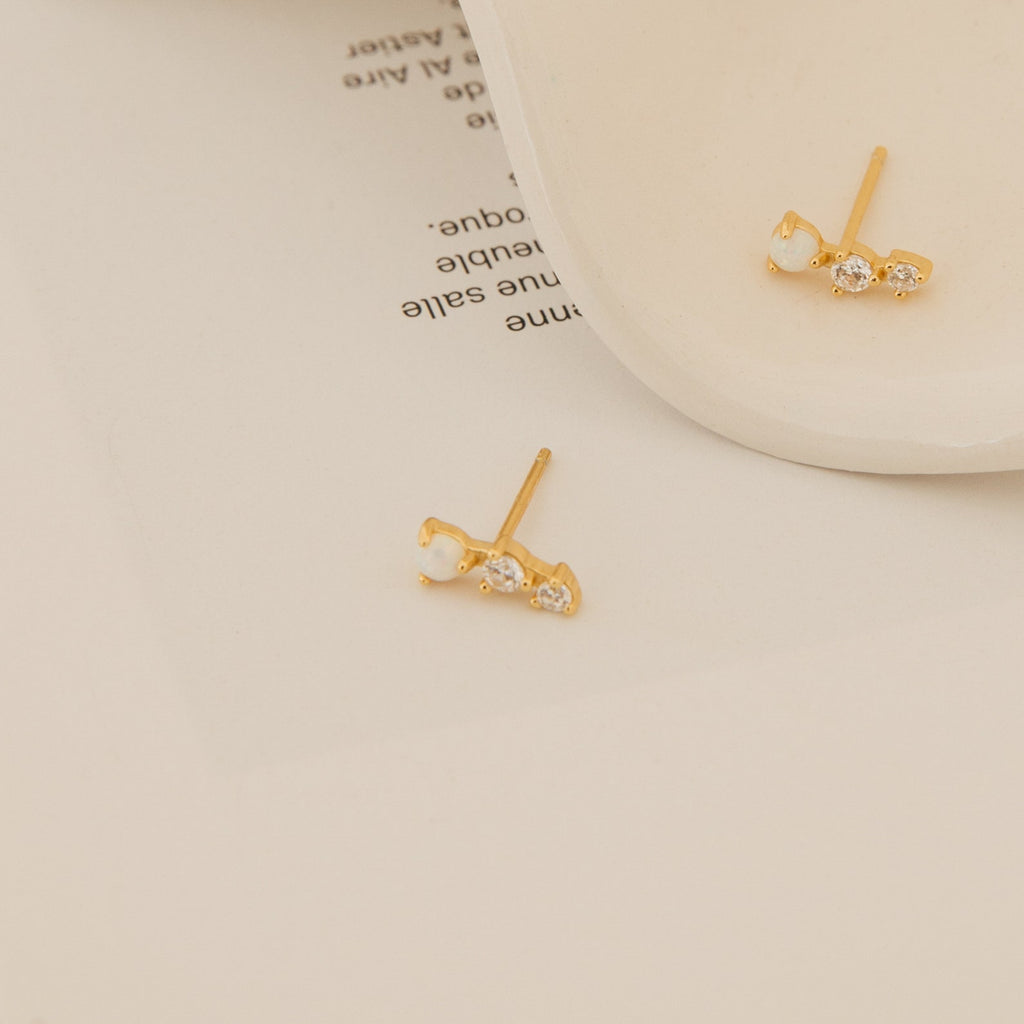 Maeve Opal Studs: Gold gemstone earrings featuring three small clear stones each, beautifully displayed on a white dish with a beige background—ideal for anyone seeking elegant statement earrings.
