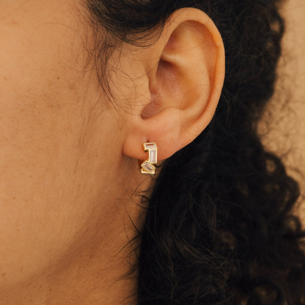 Close-up of a person's ear wearing Glacier Baguette Huggies—minimalist gold earrings featuring geometric shapes with clear and yellow stones.