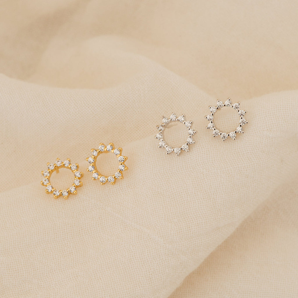 Two pairs of Helen Diamond Sun Studs—one gold, one silver—are displayed on beige fabric. Ideal for those seeking minimal, shimmery diamond earrings with a sun motif.