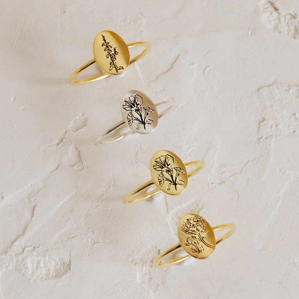 Four Flower Oval Rings—three gold, one silver—each engraved with a unique birth flower, make a beautiful personalized jewelry set displayed on a textured surface.