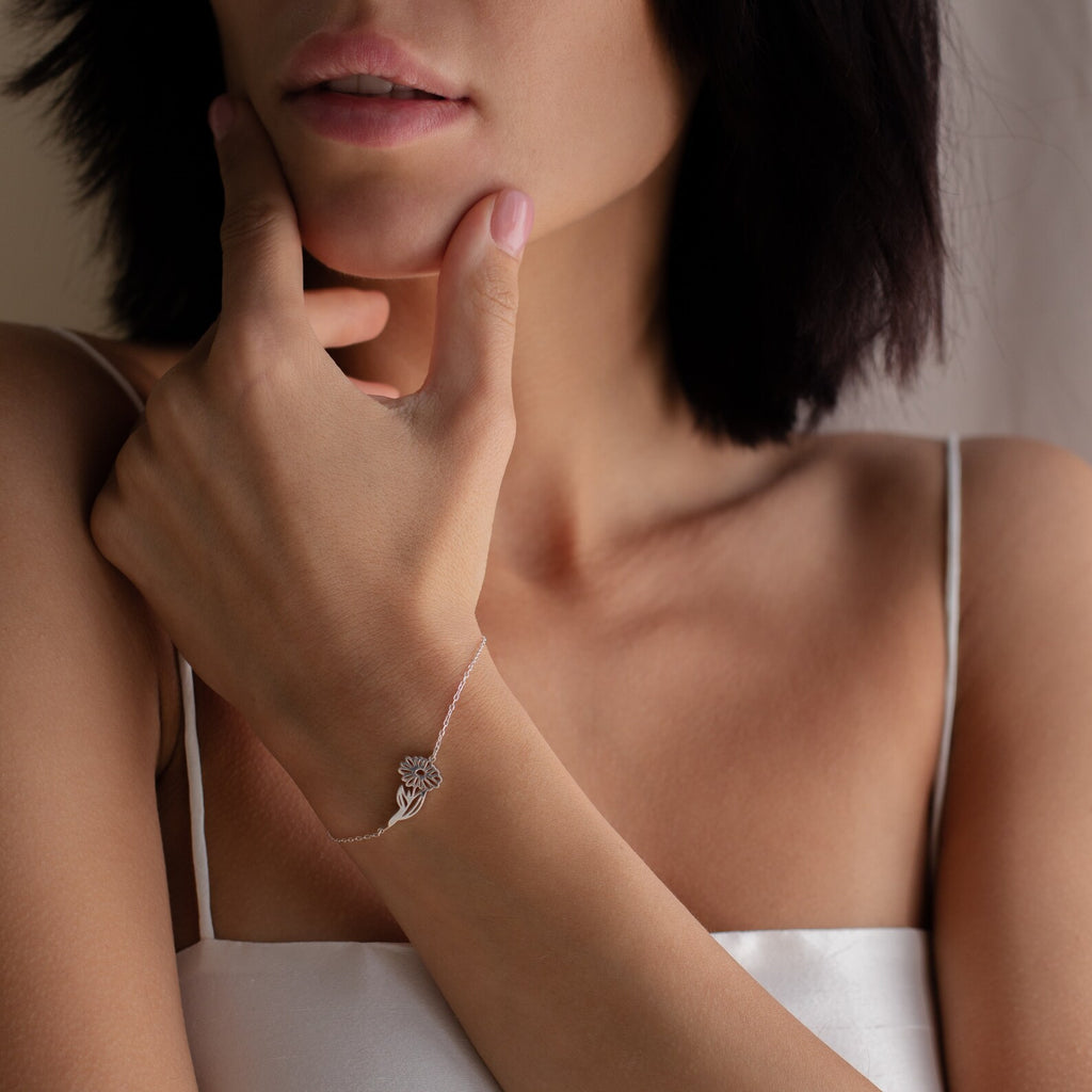 A woman in a white satin top wears a Custom Flower Bracelet in No Stone, featuring a birth flower charm, as she thoughtfully touches her chin.
