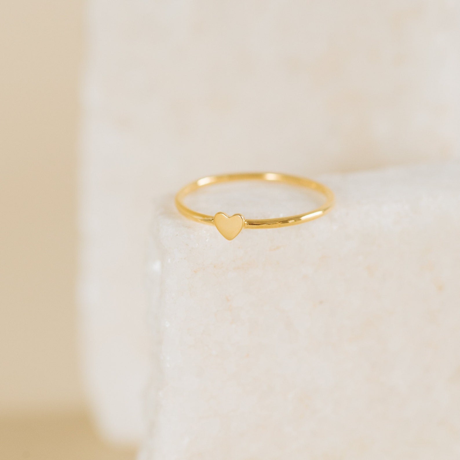 The Nala Tiny Heart Ring is a delicate gold stacking ring featuring a tiny heart-shaped centerpiece, shown on a light textured surface.