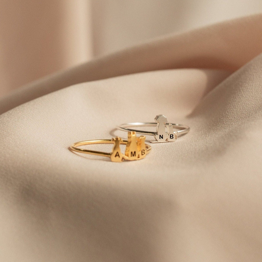 Two delicate Pet Initial Rings, one gold and one silver, with letter charms shown on soft beige fabric—a personalized jewelry piece ideal for pet lovers.