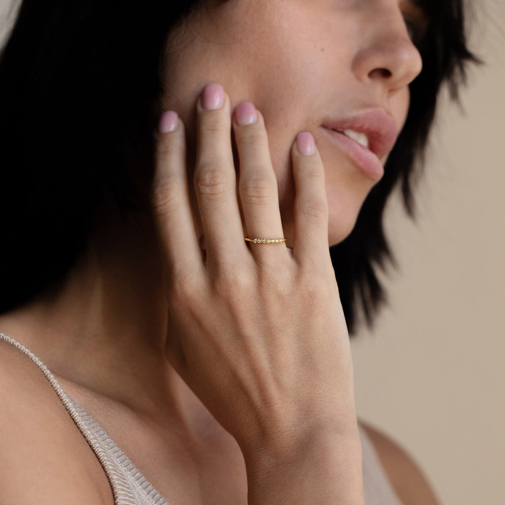 A woman with short dark hair displays the Isabelle Diamond Ring—a dainty band adorned with small diamonds—on her finger near her face.