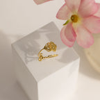 The Flower Name Ring features a gold band with a rose design and "amour" script, displayed on a white block beside a pink flower. This custom jewelry is a sentimental gift for mom, adding personalized charm to her collection.