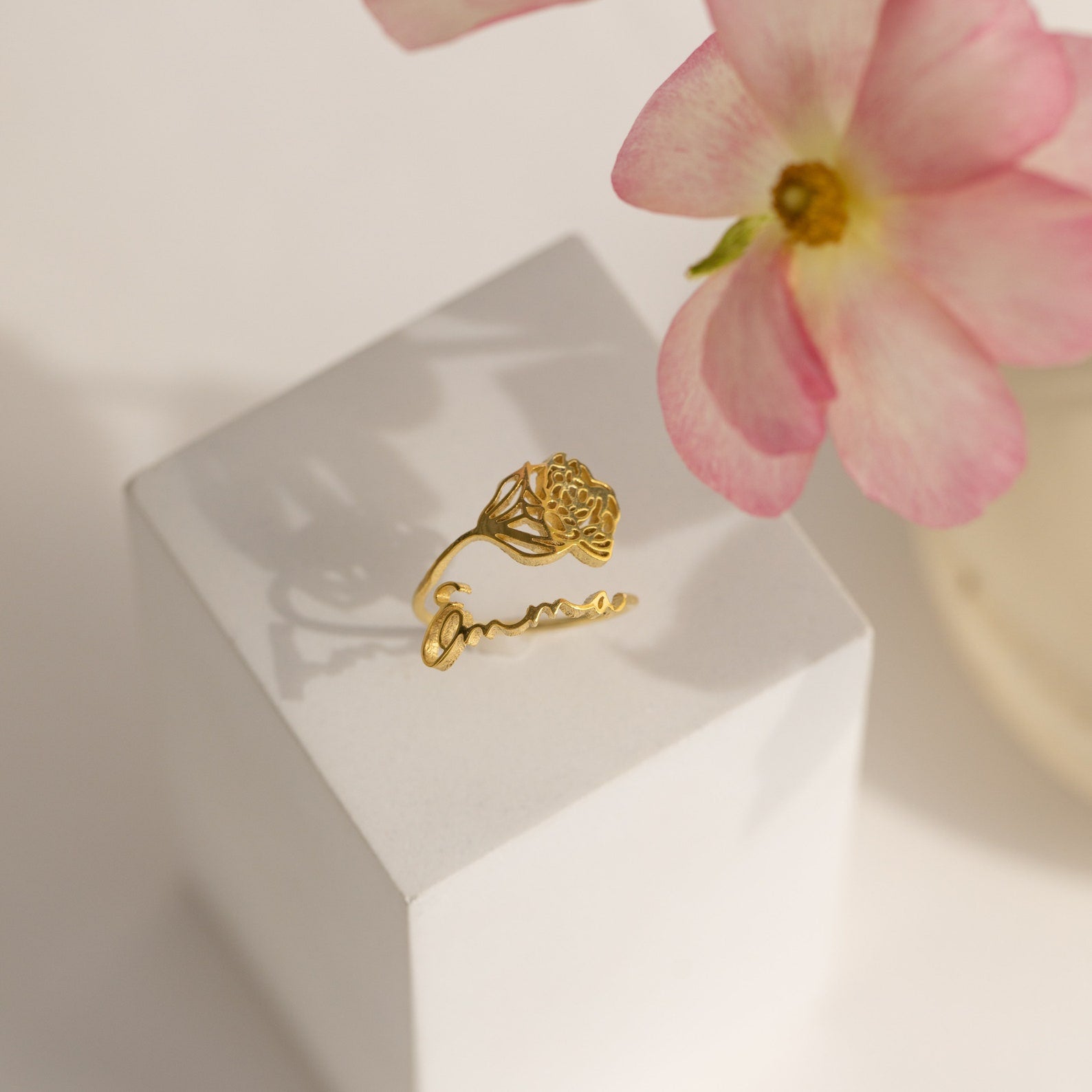 The Flower Name Ring features a gold band with a rose design and "amour" script, displayed on a white block beside a pink flower. This custom jewelry is a sentimental gift for mom, adding personalized charm to her collection.