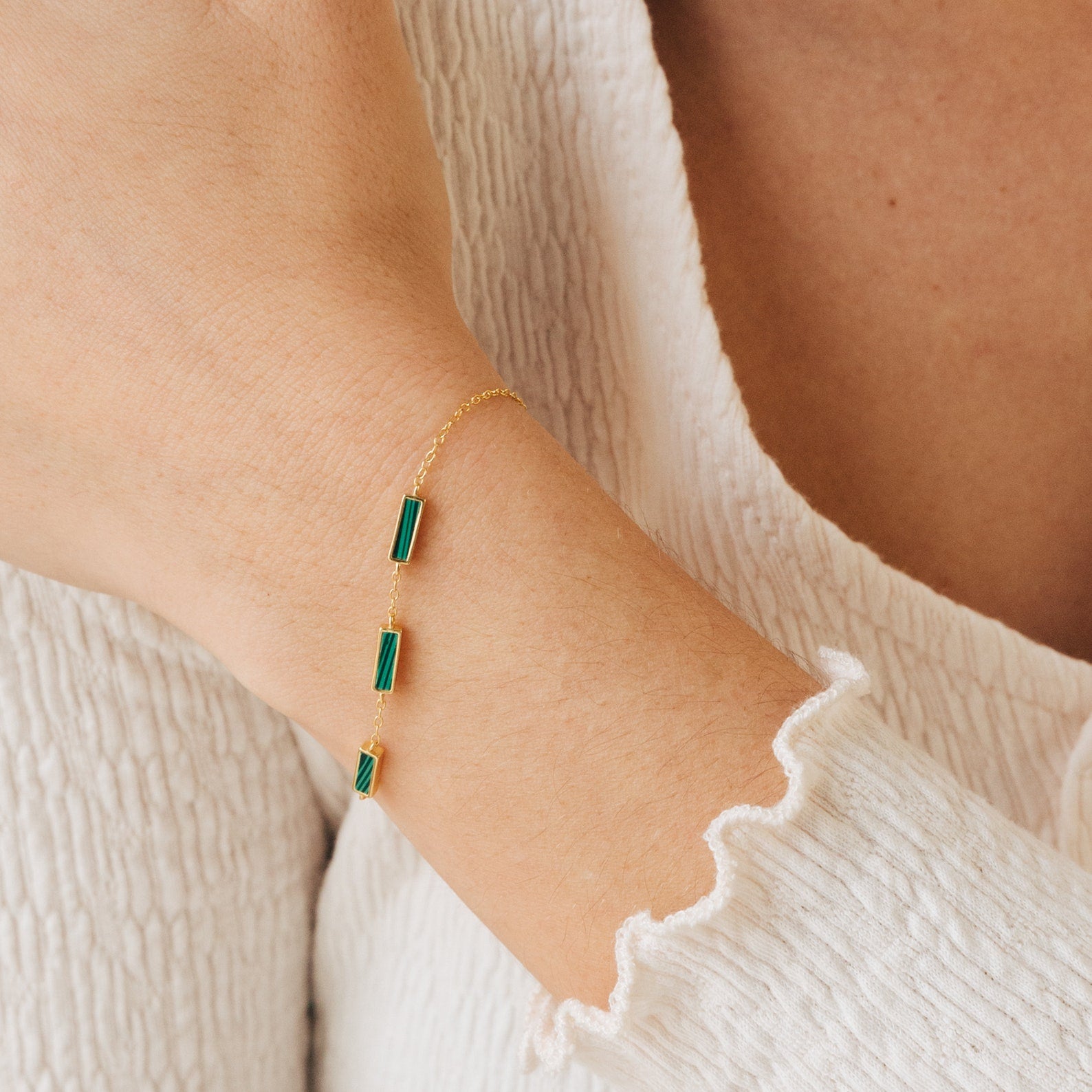 The Malachite Bar Bracelet features a gold dainty chain with three green rectangular malachite stones, shown on a wrist with a white textured top—a perfect handmade boho accessory.