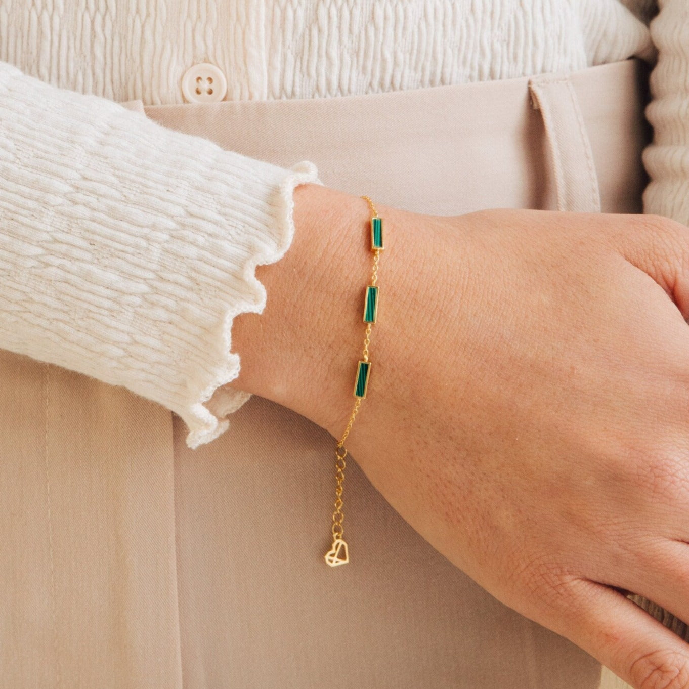 A hand models the Malachite Bar Bracelet, featuring green rectangular malachite stones and a delicate chain, styled with a cream blouse and beige pants.