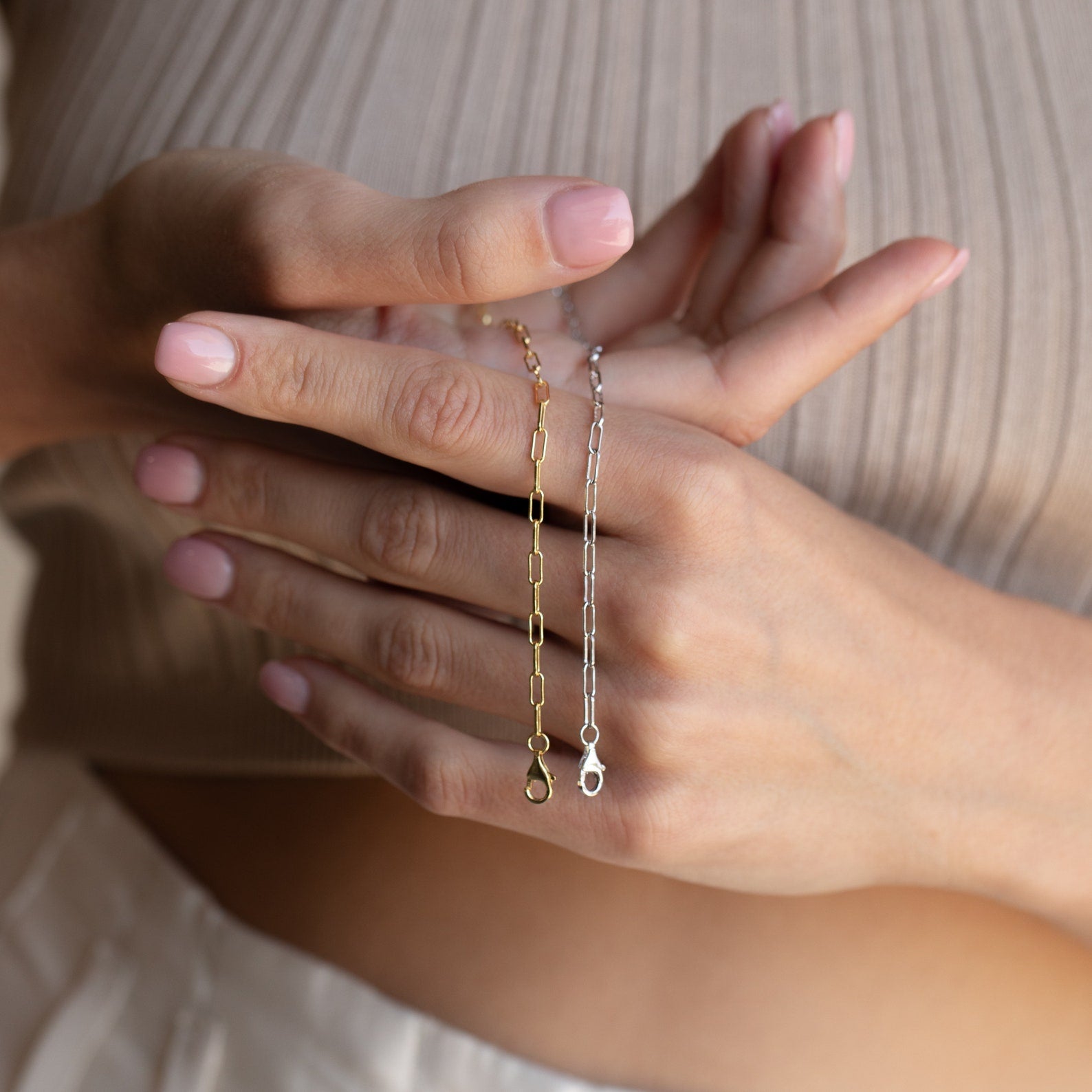 Close-up of hands with light pink nails wearing a Paperclip Chain Bracelet alongside other stacking bracelets, featuring a dainty charm, all against a ribbed beige top.