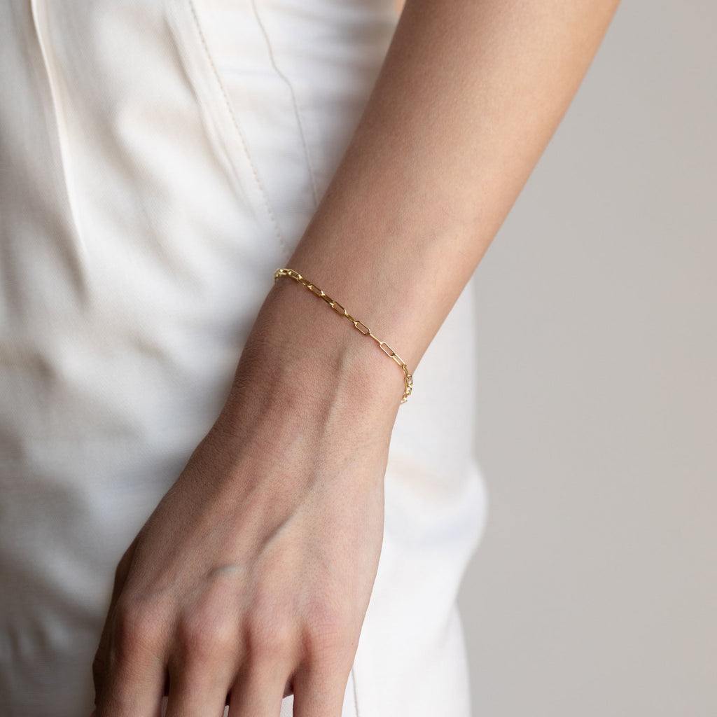 A hand wearing a dainty Paperclip Chain Bracelet rests against white pants.
