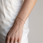 A hand wearing a dainty Paperclip Chain Bracelet rests against white pants.
