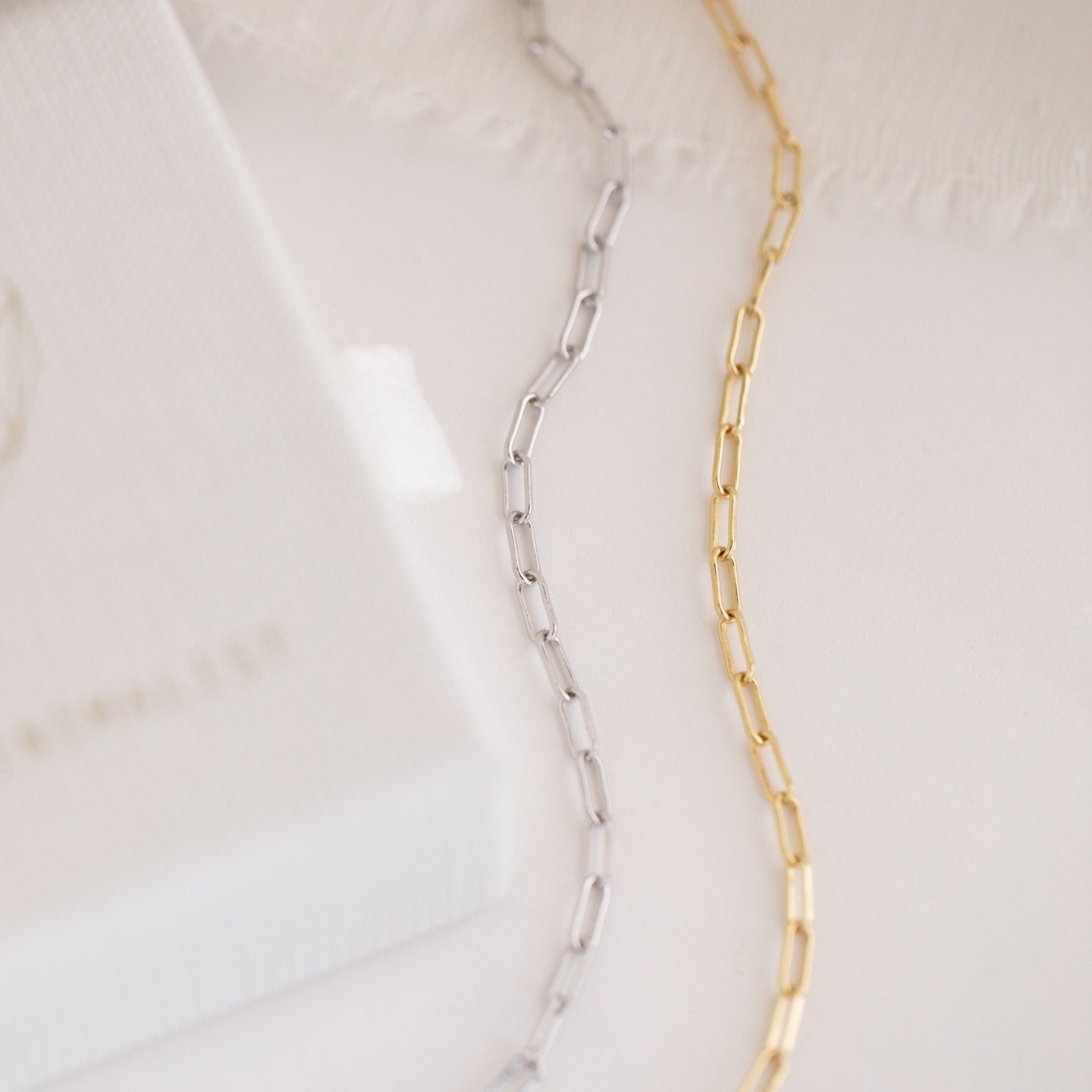 Close-up of a silver and gold Paperclip Chain Bracelet on a white surface with textured fabric in the background—perfect for stacking or pairing with your favorite dainty charms.