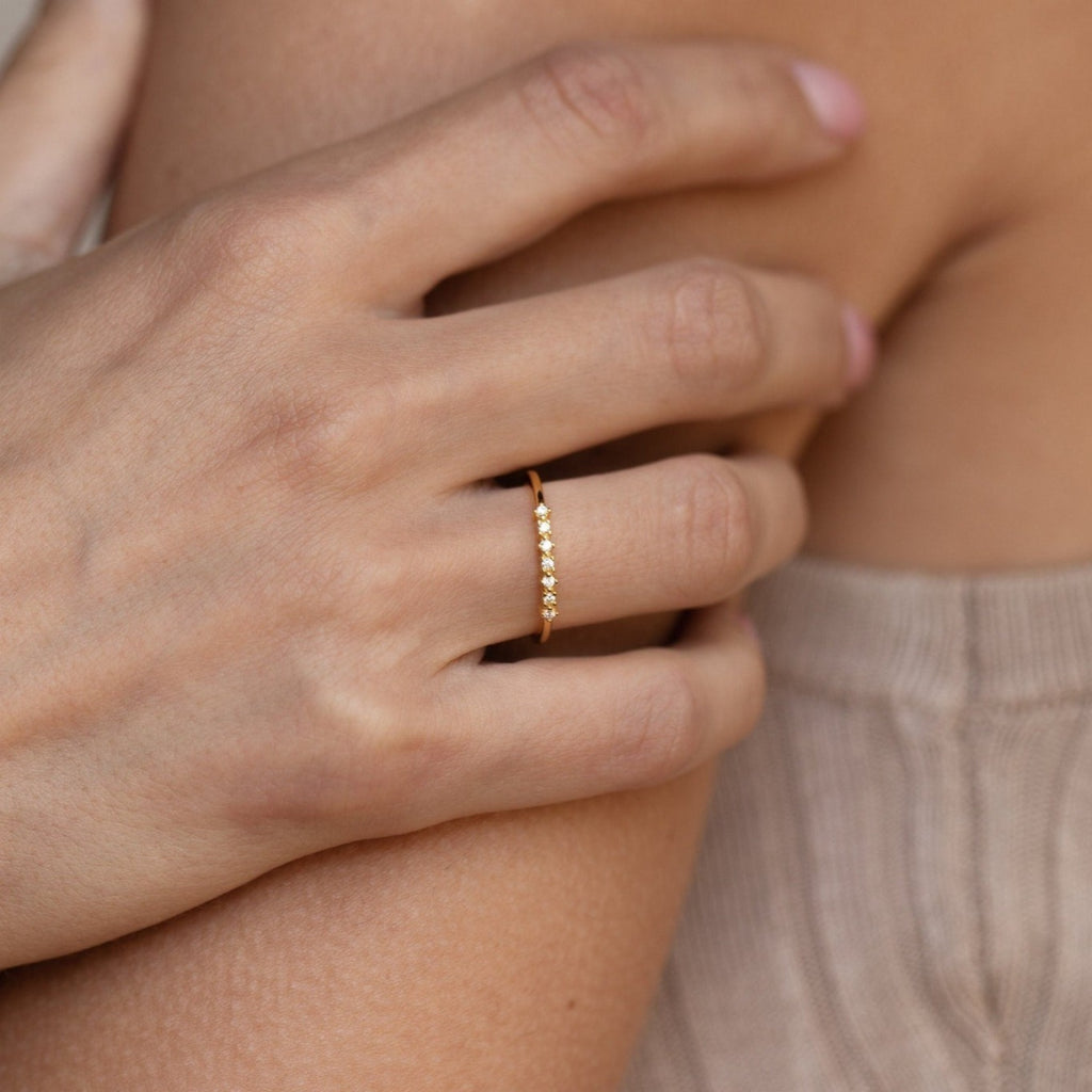 A hand wearing the Isabelle Diamond Ring, a delicate gold band with small stones, rests gently on someone’s shoulder in a beige top.