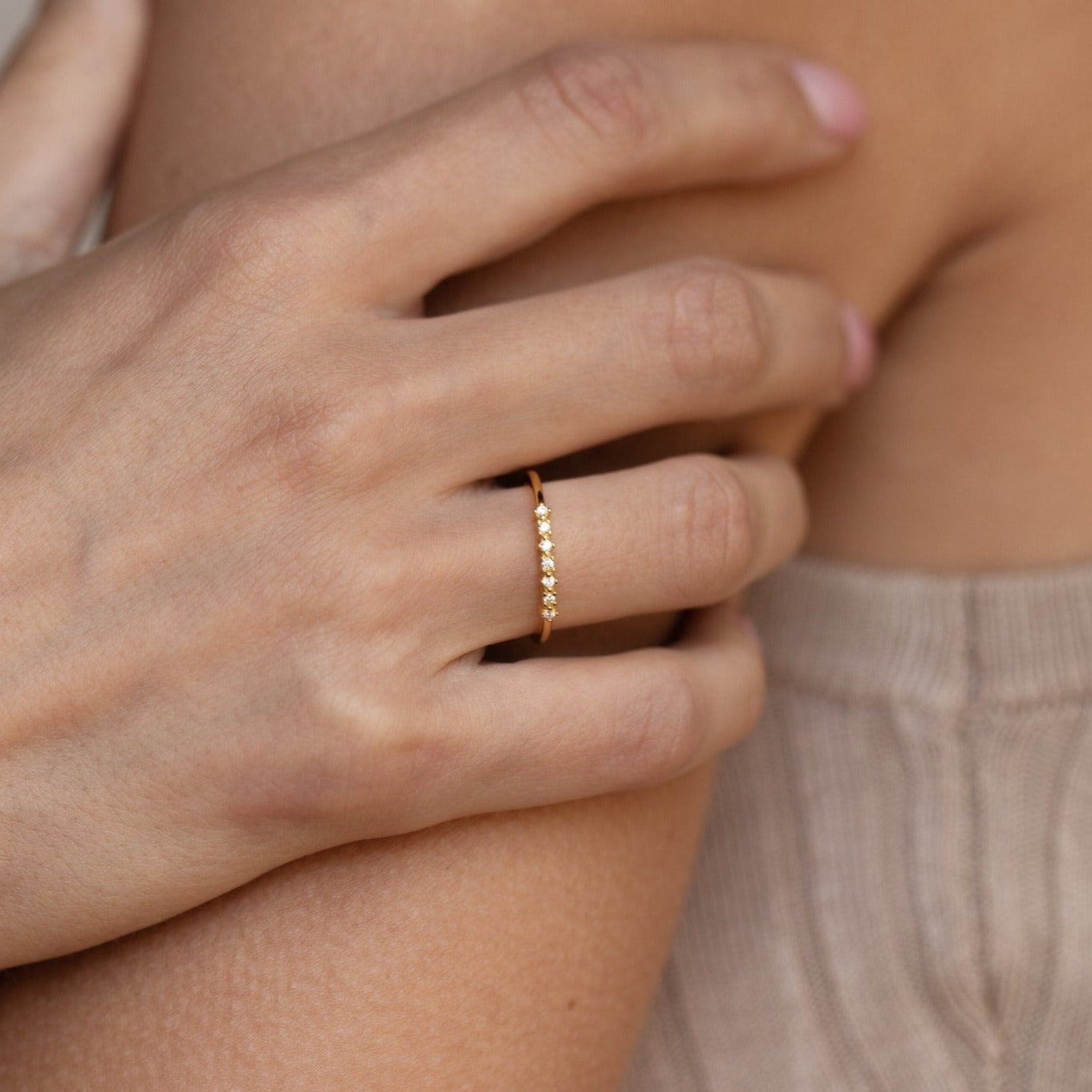 A hand wearing the Isabelle Diamond Ring, a delicate gold band with small stones, rests gently on someone’s shoulder in a beige top.
