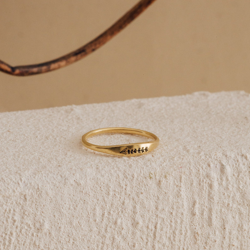 The Skinny Flower Ring in 18K Gold, with engraved symbols, rests on a textured off-white surface, showcasing the delicate elegance of botanical jewelry.