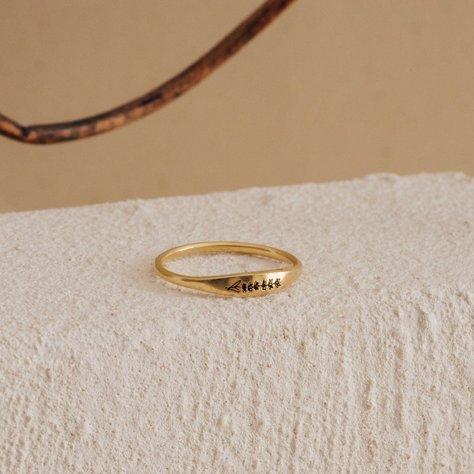 The Skinny Flower Ring in 18K Gold, with engraved symbols, rests on a textured off-white surface, showcasing the delicate elegance of botanical jewelry.