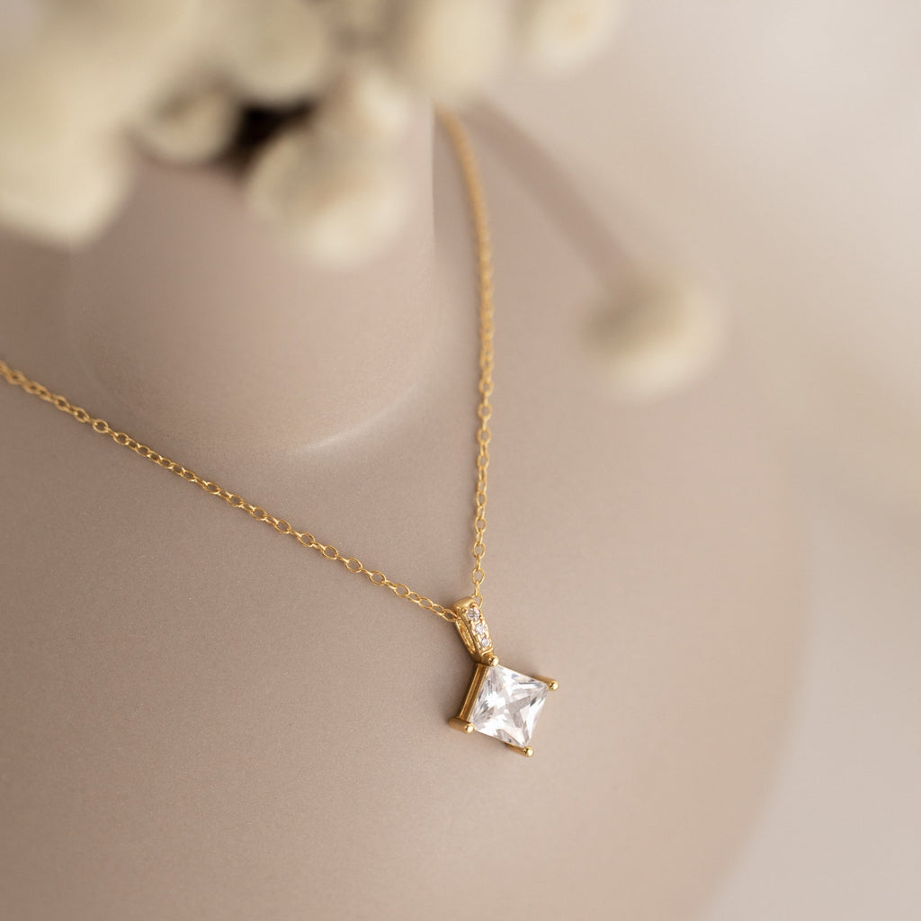 Showcased on a soft beige background, the Carina Diamond Necklace features a gold chain with a square, clear diamond pendant—experience its timeless elegance.