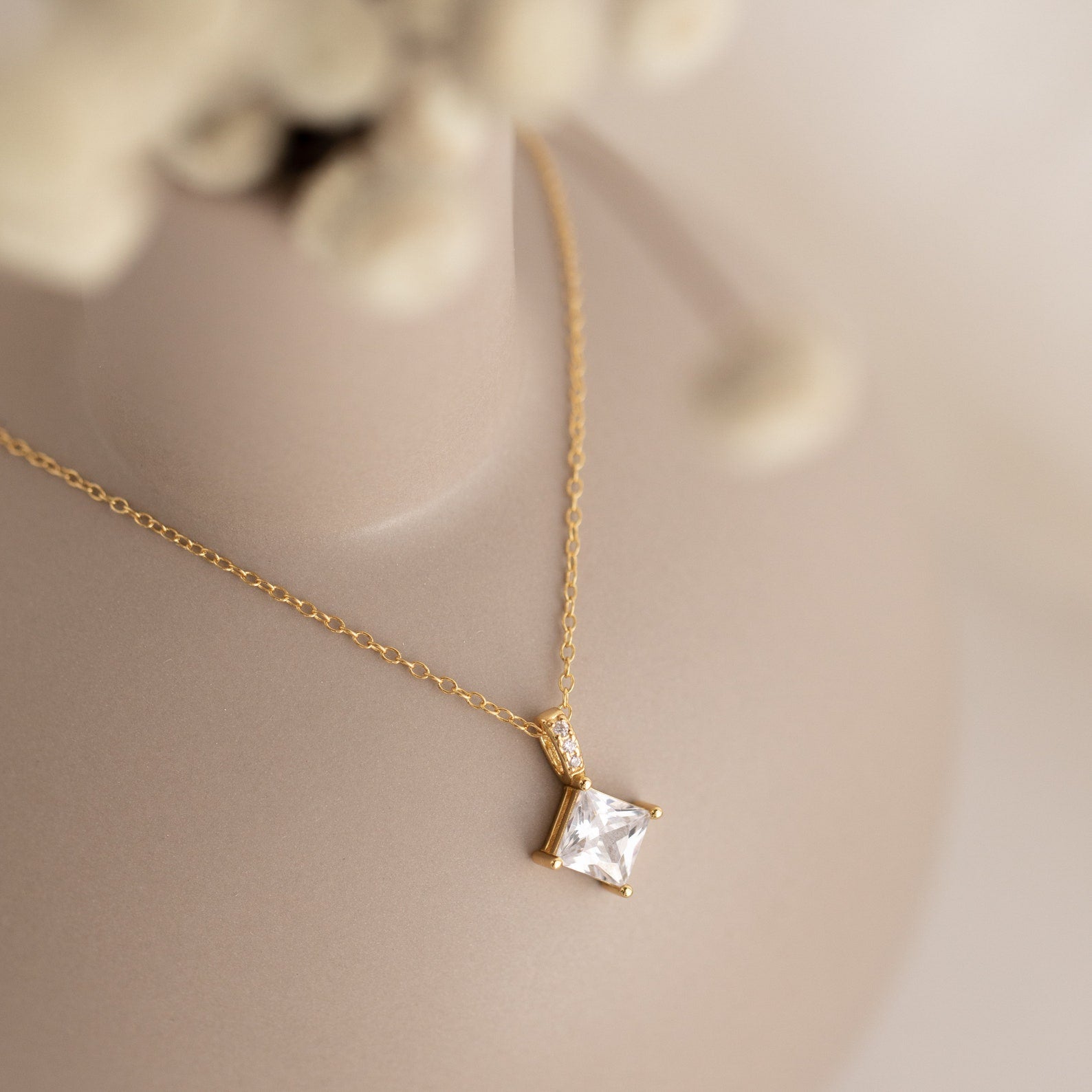 Showcased on a soft beige background, the Carina Diamond Necklace features a gold chain with a square, clear diamond pendant—experience its timeless elegance.