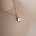 Close-up of a gold necklace featuring a square-cut clear gemstone pendant secured by four prongs, with tiny accent stones lining the bail, displayed on a neutral-toned bust.