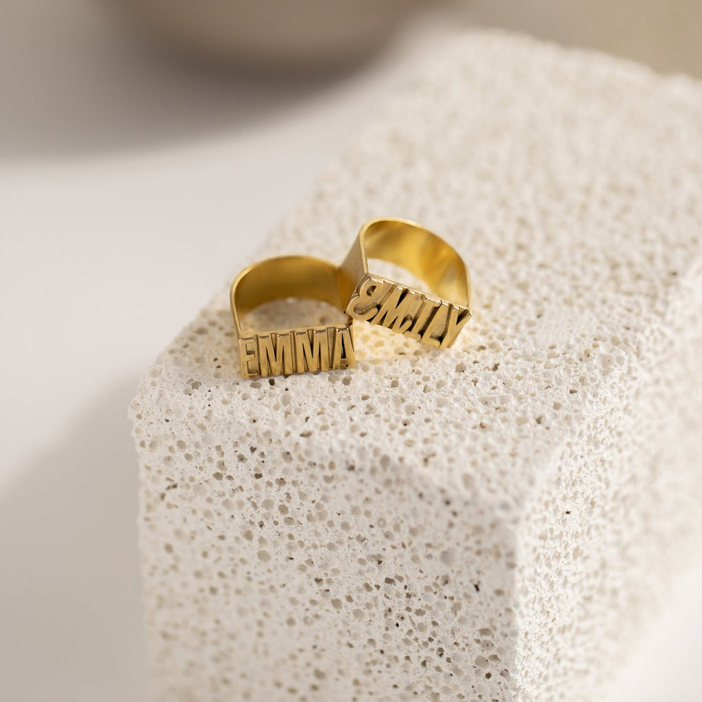 Two Bold Name Rings with "EMMA" are displayed on a textured white stone block, highlighting a unique personalized ring style.