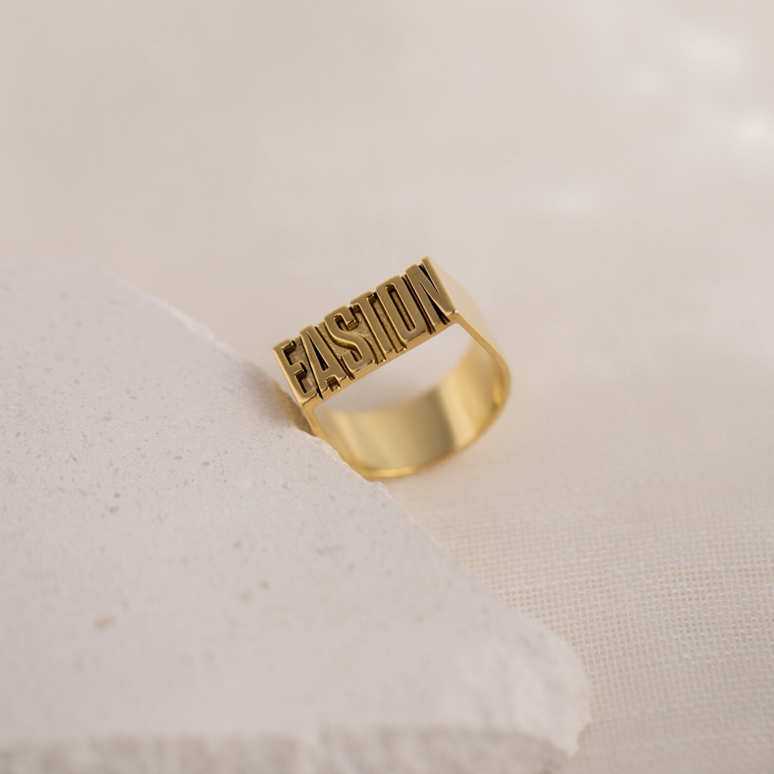 The Bold Name Ring, a gold statement piece featuring the word EASTON in bold letters, displayed on a textured, light-colored surface.