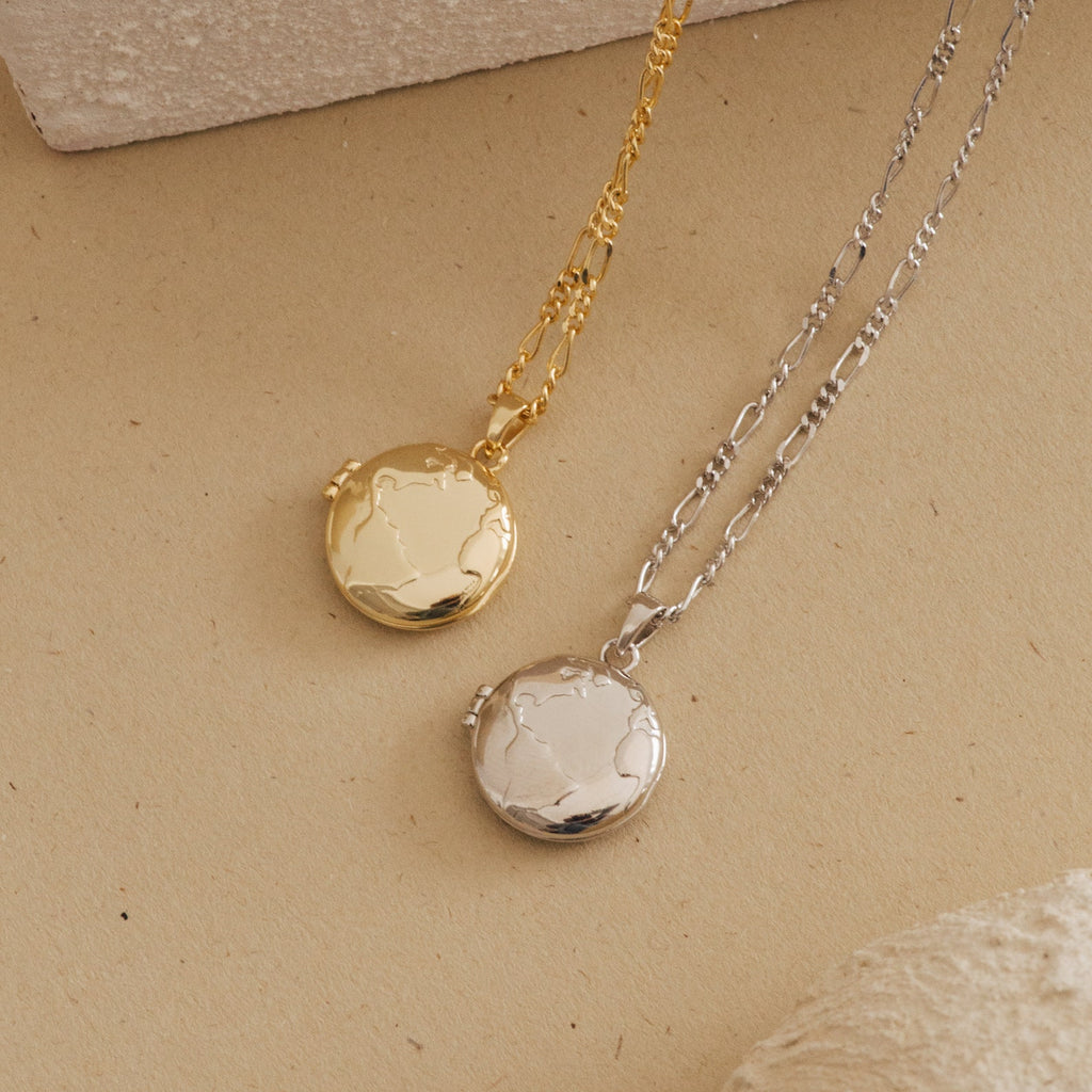 Flat lay of two globe-engraved locket necklaces, one in polished gold and the other in polished silver, both with textured chain links, displayed on a beige textured surface.