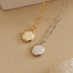 Flat lay of two globe-engraved locket necklaces, one in polished gold and the other in polished silver, both with textured chain links, displayed on a beige textured surface.